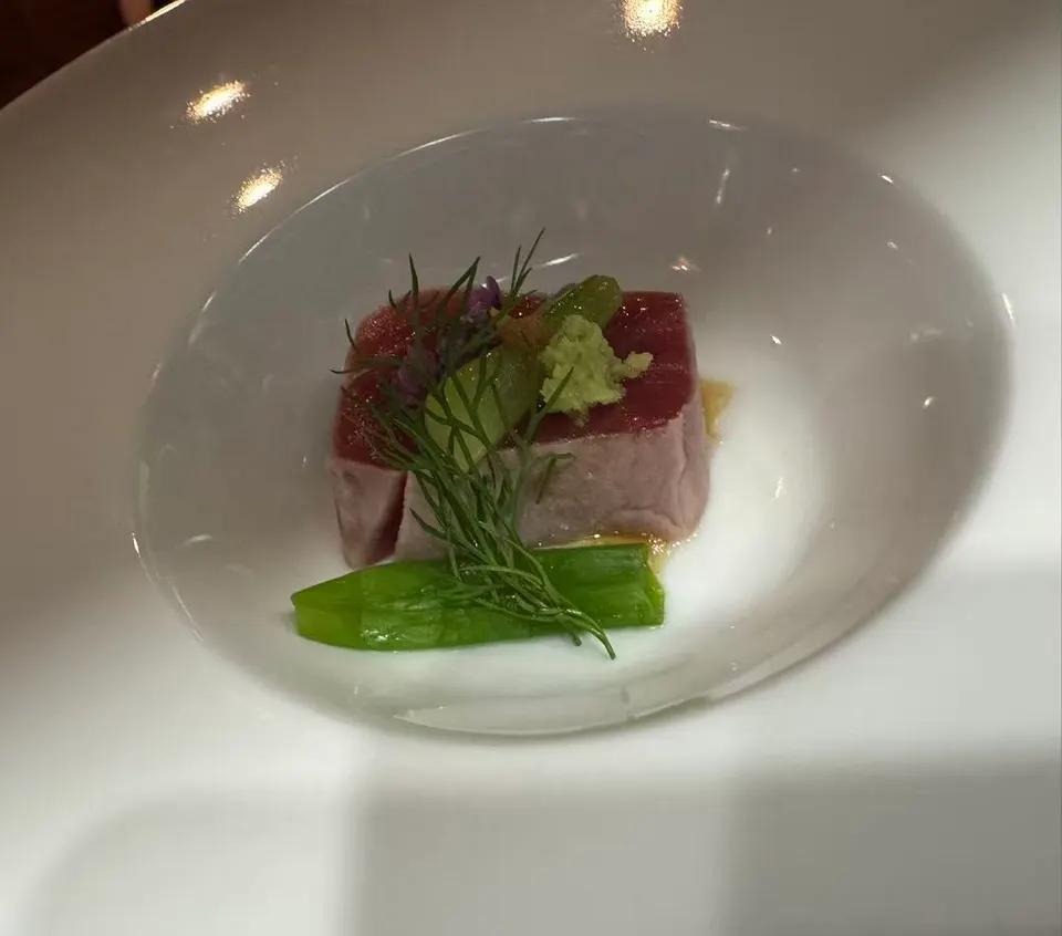 A single piece of seared tuna sashimi served in a white bowl, garnished with fresh dill, a green vegetable, and a dollop of wasabi.