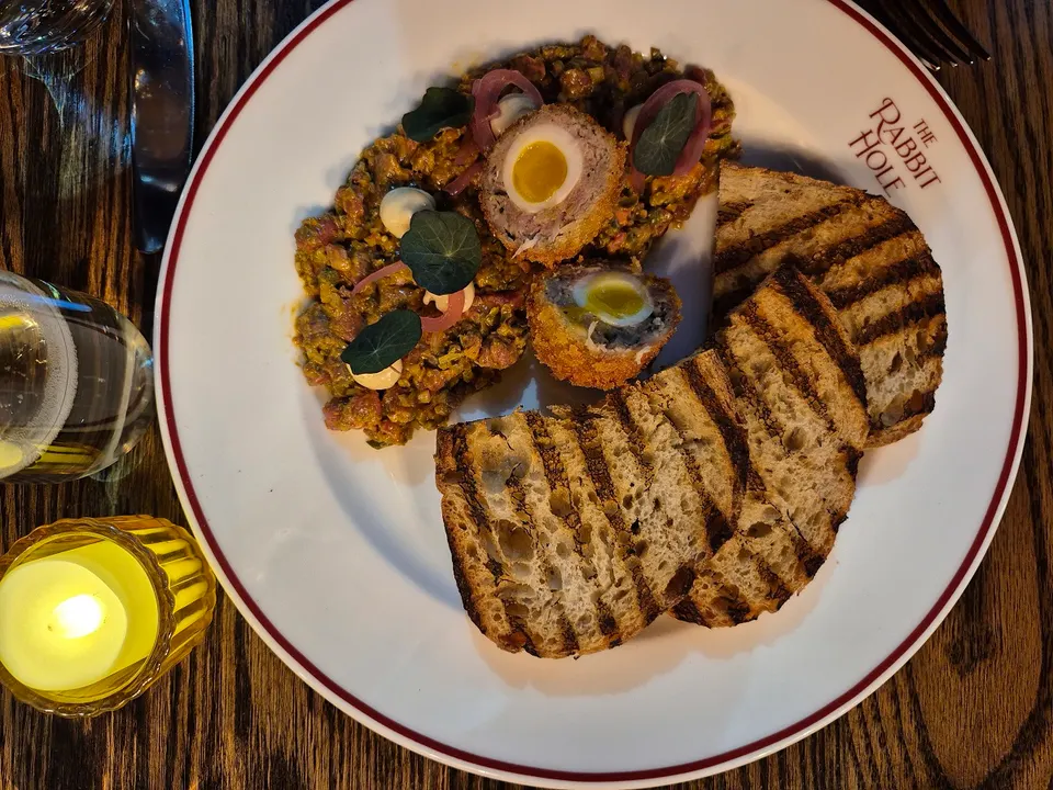 A plate of beef tartare topped with halved scotch eggs, served with grilled sourdough bread slices, on a white plate with a red rim and 'The Rabbit Hole' logo.