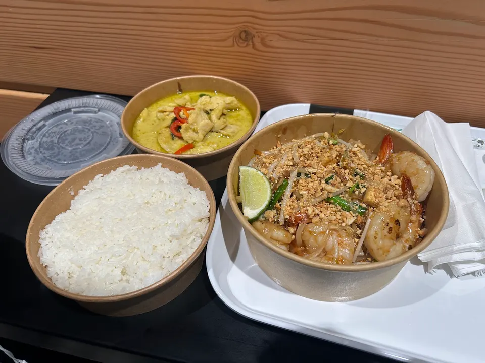 A spread of Thai takeout dishes from Kao Kang by Koh Lipe, featuring a bowl of white rice, green curry with chicken, and Pad Thai with shrimp, crushed peanuts, and a lime wedge.