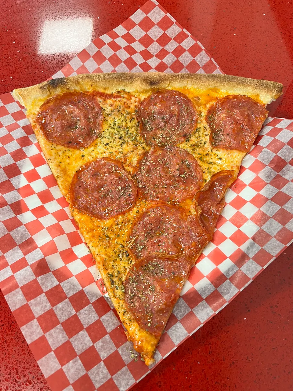 A large slice of pepperoni pizza seasoned with dried herbs, served on red and white checkered wax paper over a red countertop.