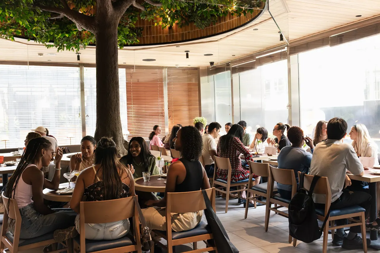 A busy indoor dining area at Everyside - Social Eatery & Taphouse, filled with patrons sitting at wooden tables under a large decorative indoor tree with lush green leaves.