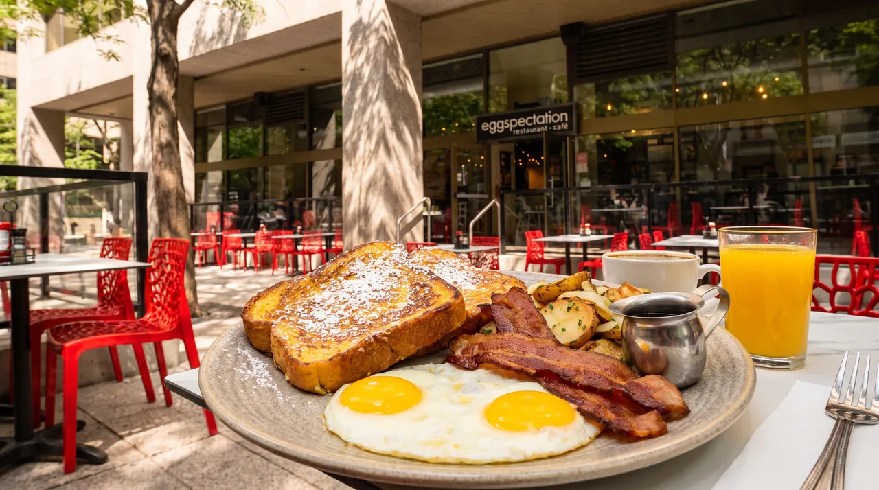 A breakfast platter with French toast, two sunny-side-up eggs, bacon, and home fries, served with orange juice and coffee on an outdoor patio at eggspectation.