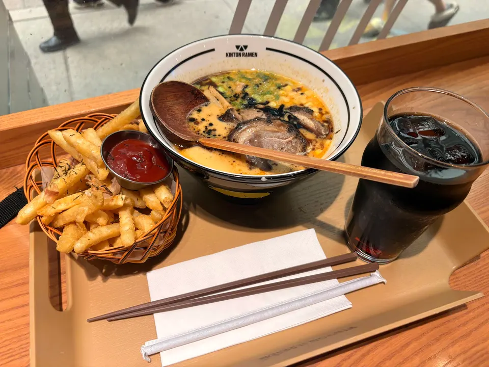 A tray containing a bowl of ramen with pork slices, a basket of french fries with ketchup, and a glass of iced dark beverage with chopsticks and a straw.