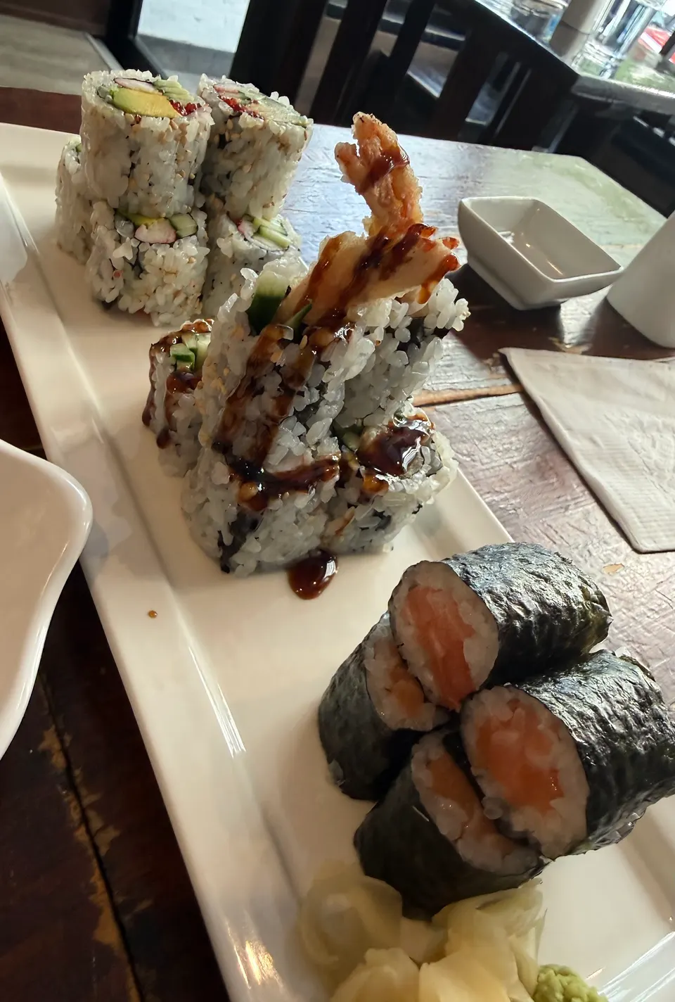 A white rectangular platter on a wooden table holds three different types of sushi rolls, including salmon maki, a tempura roll with dark sauce, and California rolls, served with ginger and wasabi.