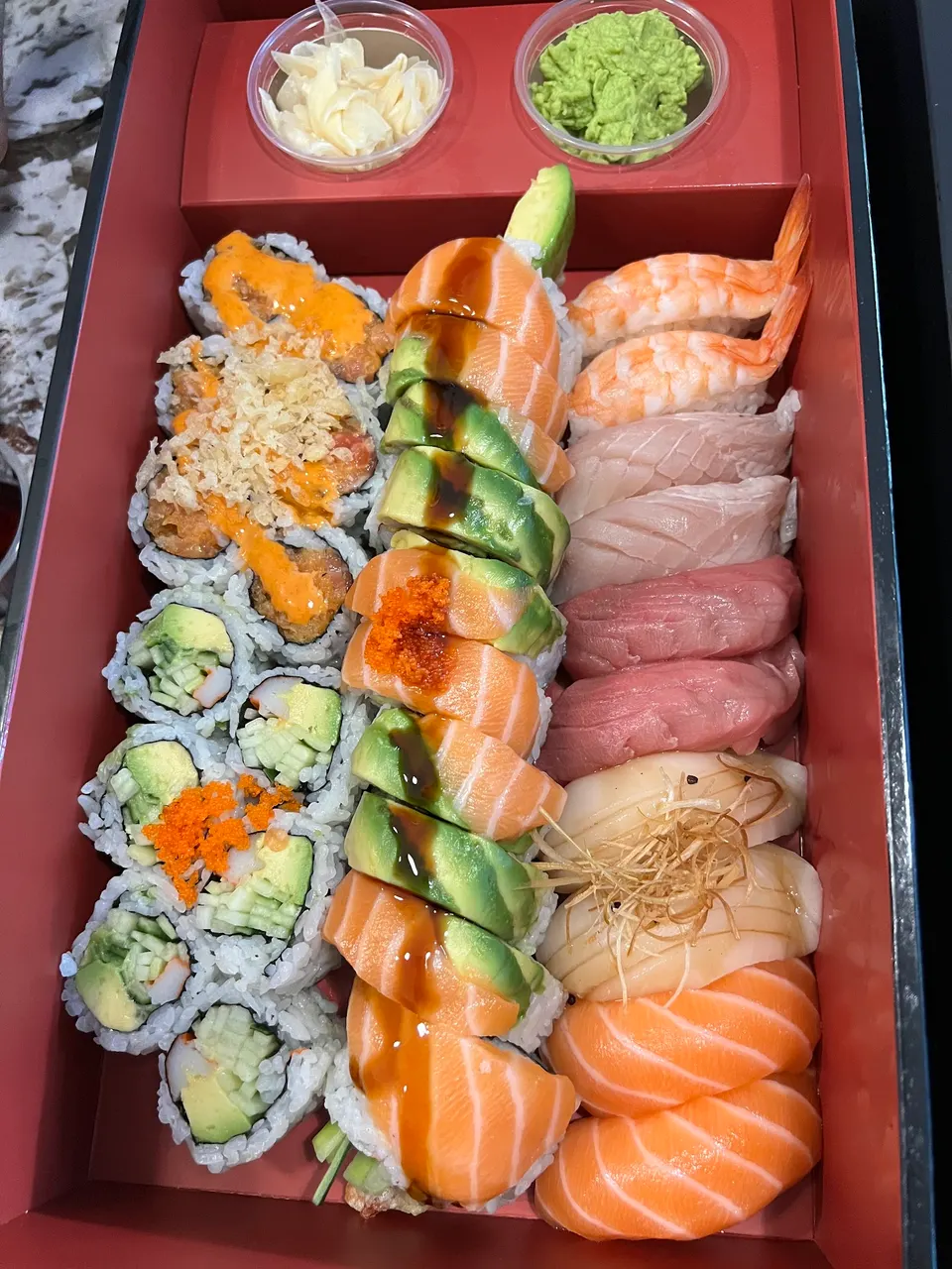 A red bento box filled with an assortment of sushi, including a dragon roll topped with salmon and avocado, spicy tuna rolls, California rolls, and various nigiri like shrimp, tuna, and salmon, served with ginger and wasabi.