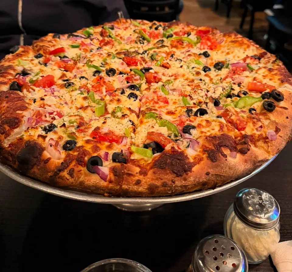 A large, freshly baked gourmet pizza loaded with melted cheese, green bell peppers, red onions, black olives, and diced tomatoes, served on a metal stand next to shaker bottles.