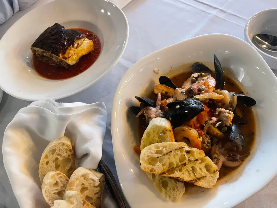 A table setting at Volos Greek Cuisine featuring a bowl of seafood stew with mussels, shrimp, and grilled bread, a plate of baked moussaka in tomato sauce, and a basket of sliced bread.