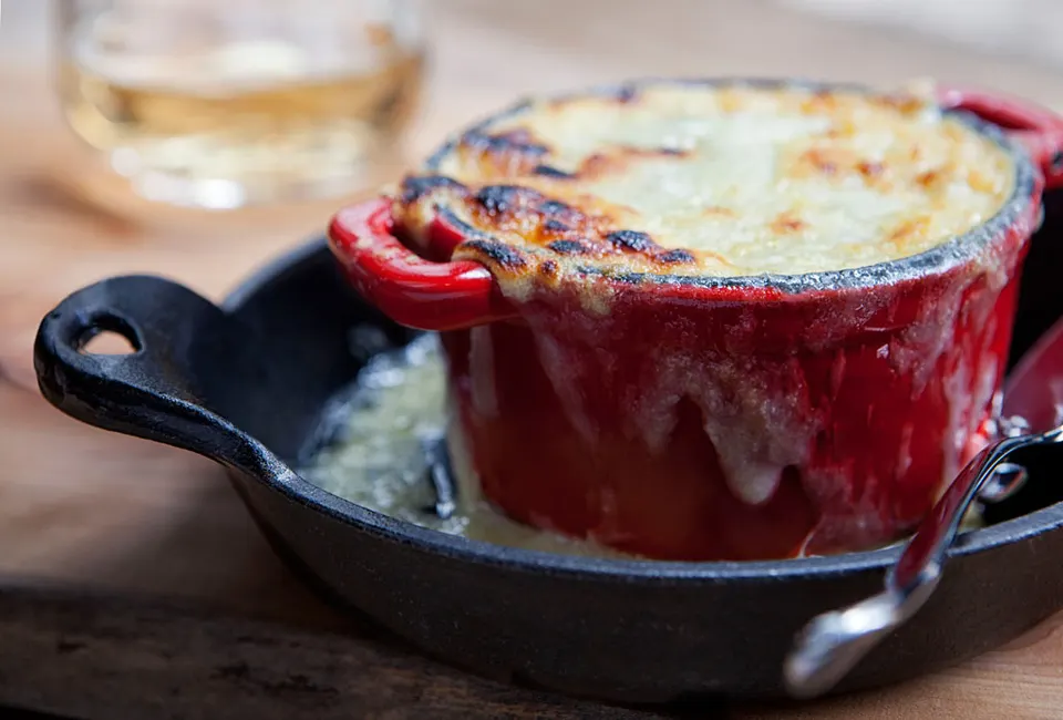 French onion soup topped with melted, browned gruyere cheese bubbling over the sides of a small red ceramic cocotte, served inside a black cast iron skillet.