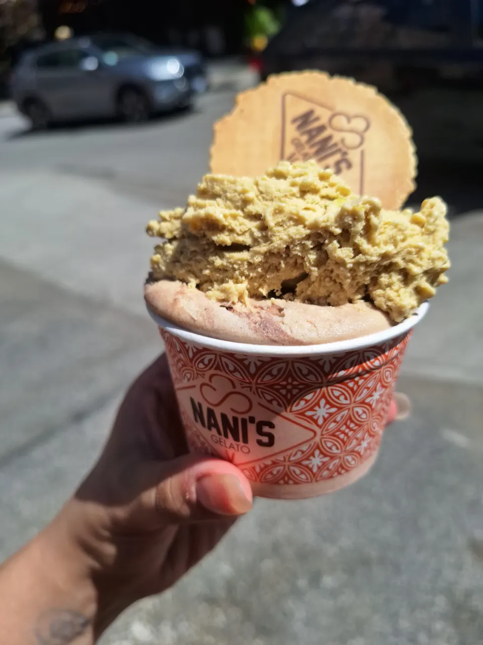 A hand holding a paper cup of Nani's Gelato with a scoop of light brown gelato topped with a scoop of pistachio gelato and a round wafer cookie.