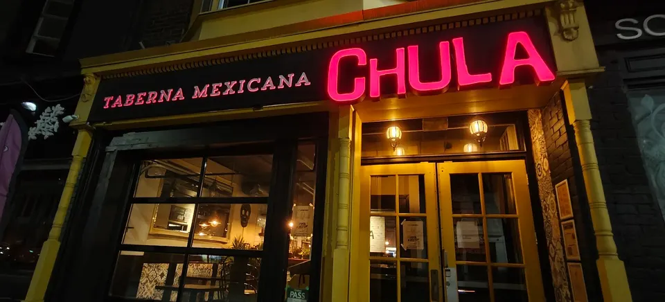 The storefront of Chula Taberna Mexicana at night, featuring a glowing pink neon sign that reads 'CHULA' and smaller pink text 'TABERNA MEXICANA' on a dark background above yellow-painted wooden doors and windows.