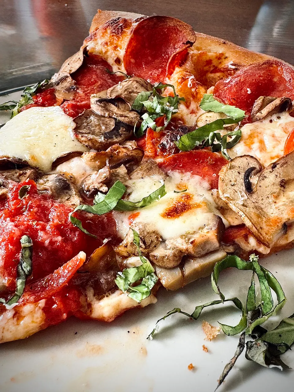 A close-up shot of a thick-crust pizza slice topped with pepperoni, sliced mushrooms, melted mozzarella cheese, rich tomato sauce, and fresh basil leaves.