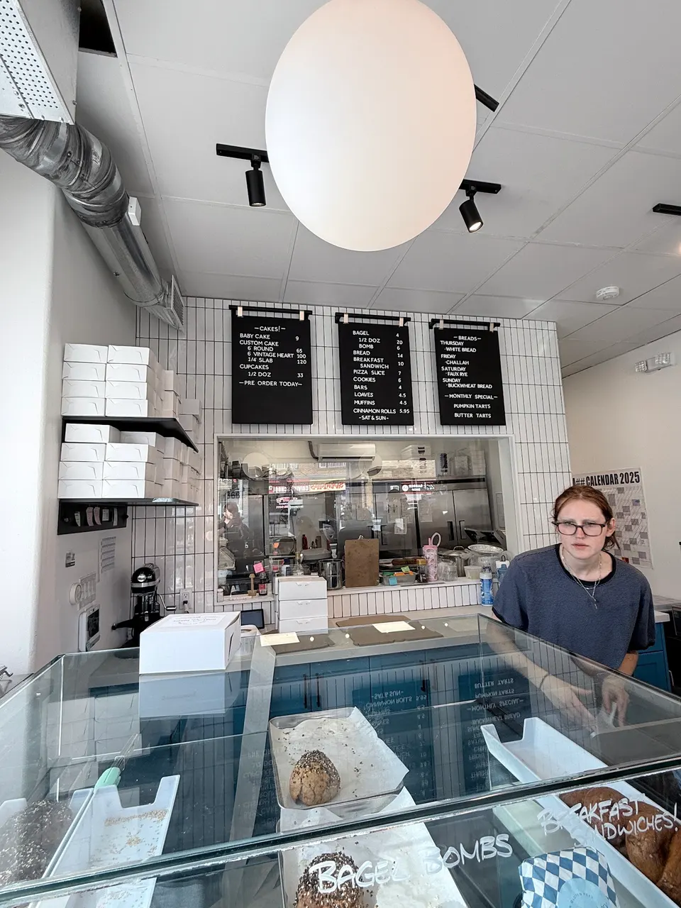 Interior of Wild Blue Bakery showing a glass display case with baked goods, a staff member behind the counter, and menu boards on a white tiled wall.