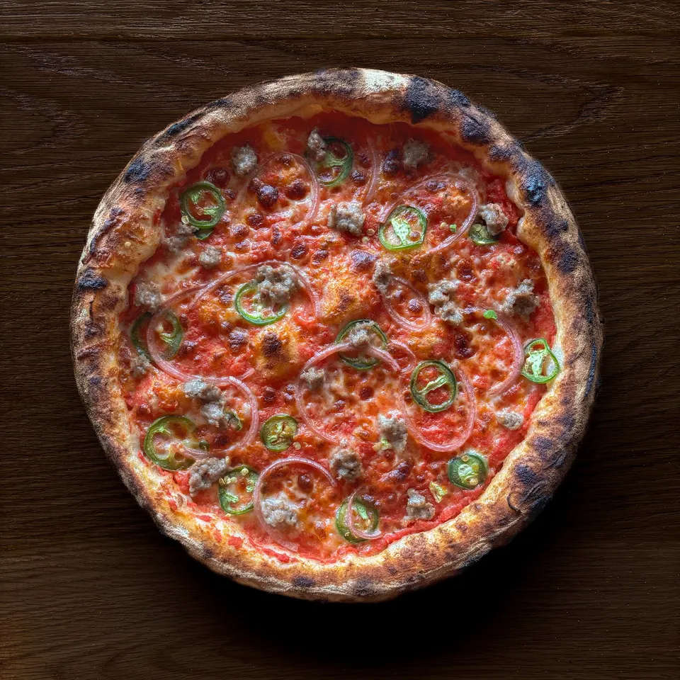 A freshly baked wood-fired pizza with a charred crust, topped with tomato sauce, melted cheese, sliced green jalapeños, red onions, and sausage chunks, centered on a dark wooden table.