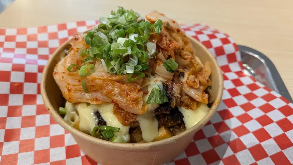 A loaded bowl filled with fries, melted cheese sauce, kimchi, and a generous topping of chopped green onions, served in a paper bowl on a red and white checkered liner.