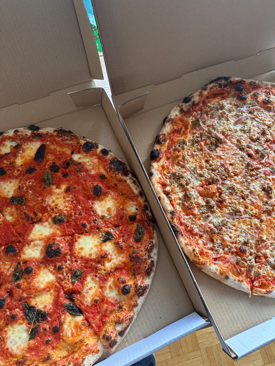 Two large, freshly baked wood-fired pizzas in open cardboard delivery boxes. One is a classic Margherita with fresh mozzarella and basil, and the other features finely crumbled sausage and red onion toppings.