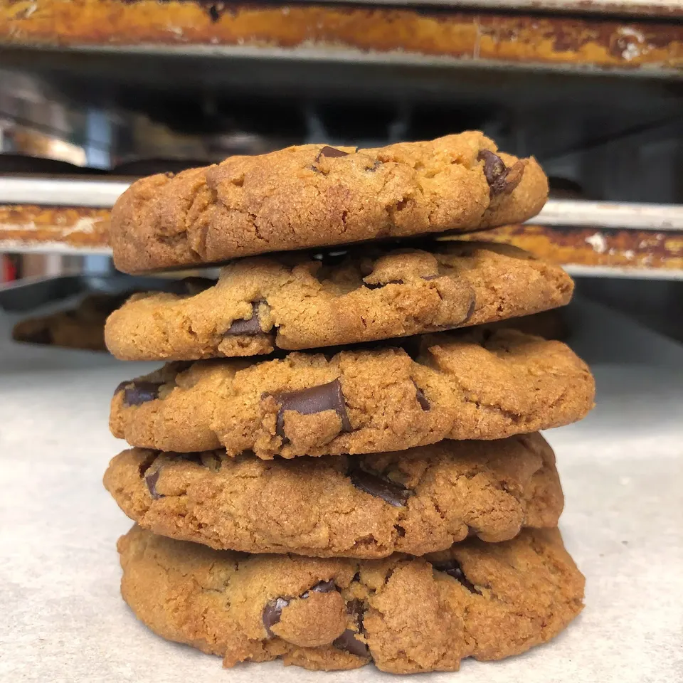 A stack of five thick, golden-brown chocolate chip cookies on a light-colored counter, with bakery racks visible in the background.
