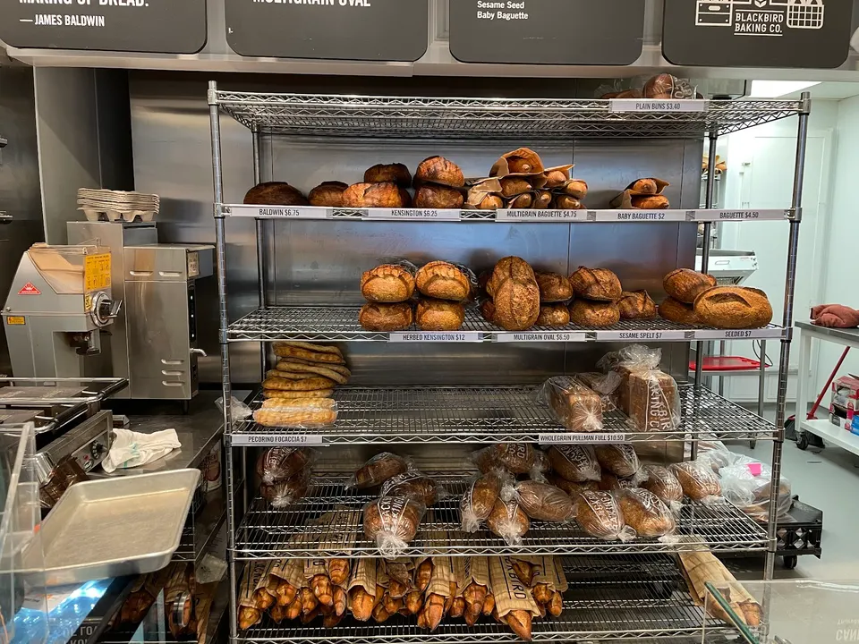 A metal wire baker's rack filled with various freshly baked breads, including loaves, baguettes, and buns, with price tags on the shelves at Blackbird Baking Co.