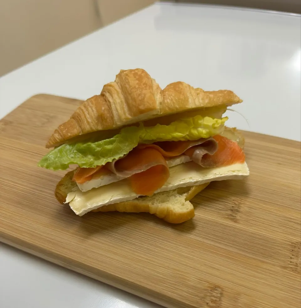 A croissant sandwich filled with brie cheese, smoked salmon, and green lettuce, served on a wooden cutting board.