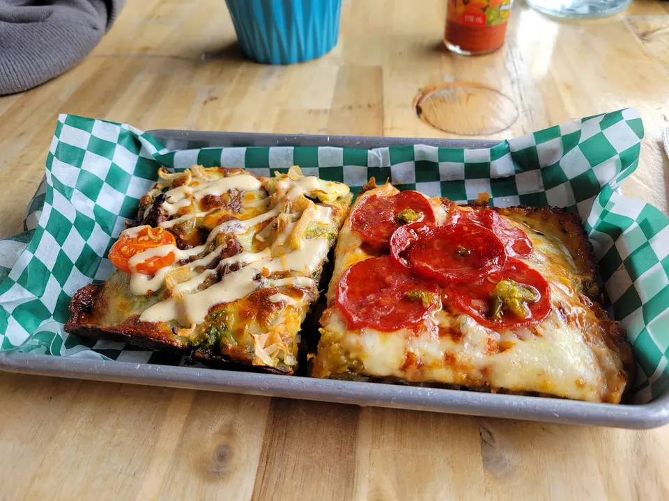 Two rectangular slices of Detroit-style deep dish pizza served side-by-side in a metal tray lined with green and white checkered paper on a light wooden table.