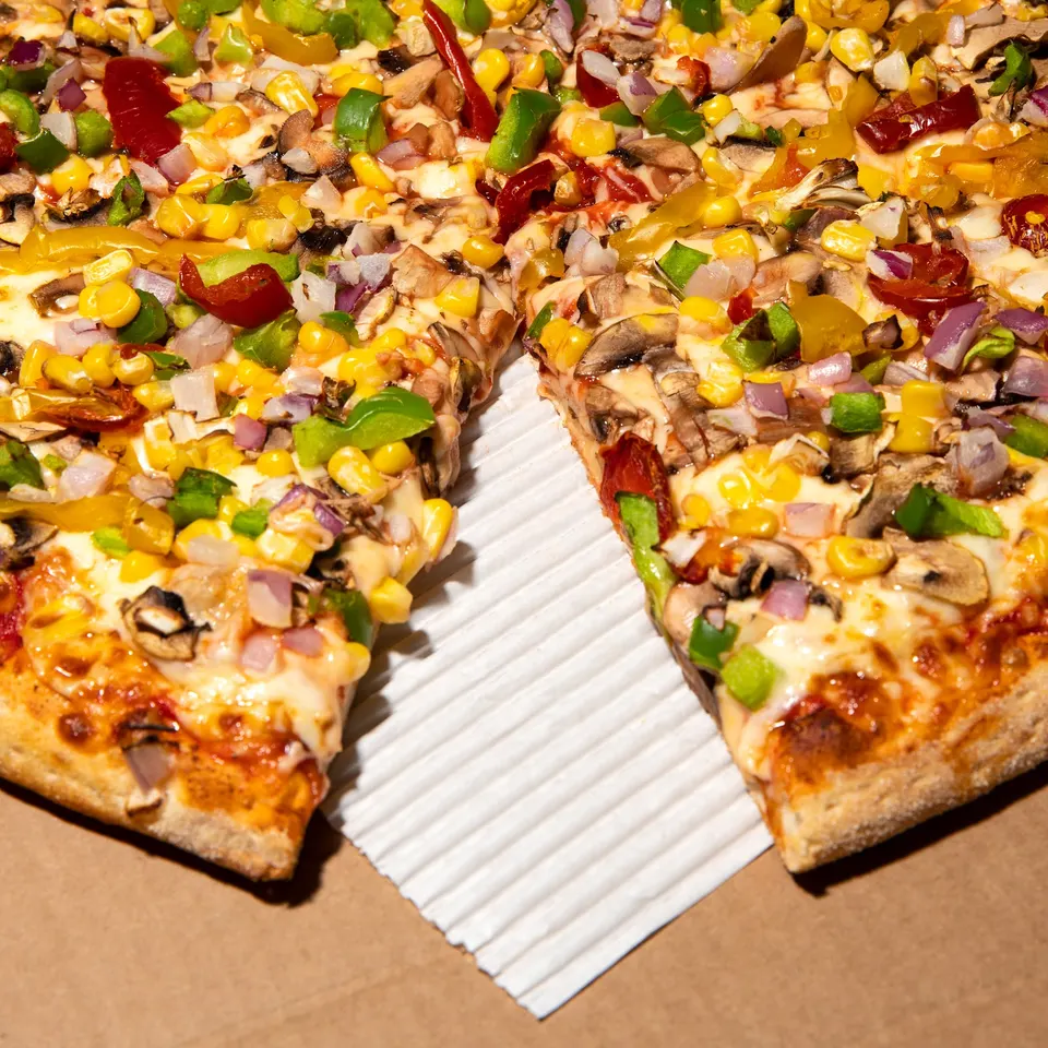 A close-up shot of a freshly baked vegetarian pizza with a slice removed, showing toppings like sweet corn, green peppers, red onions, mushrooms, and melted cheese on a thick crust.