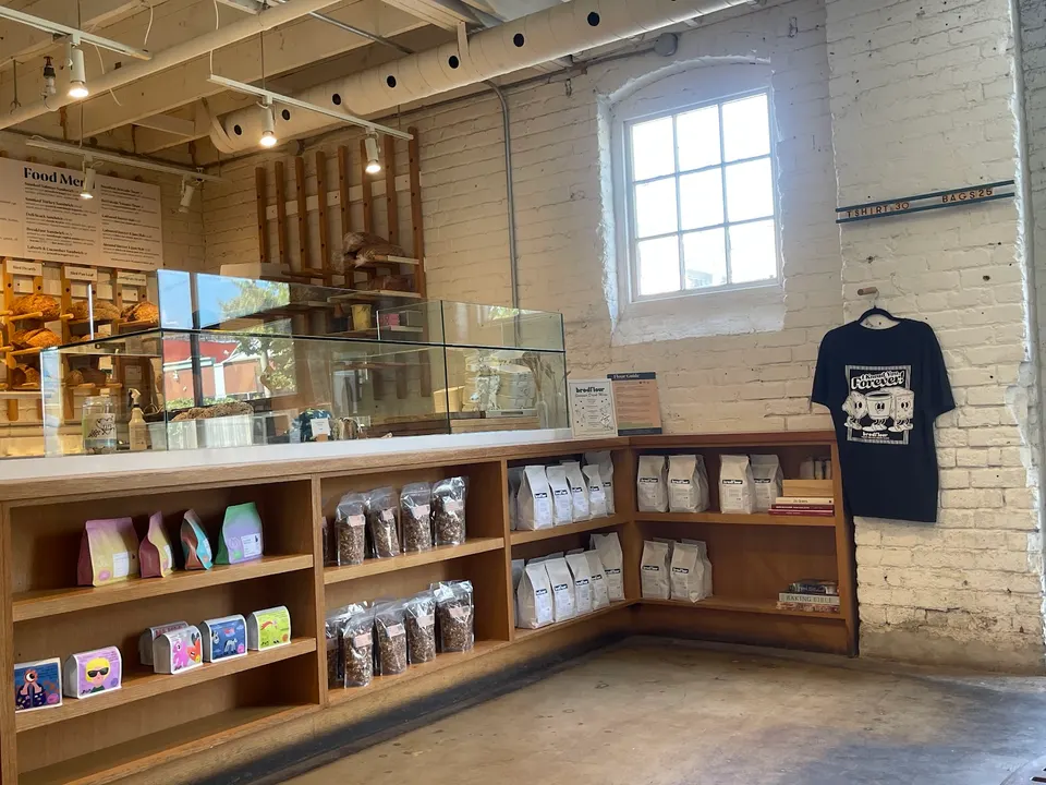 A wooden retail counter at Brodflour bakery displaying packaged coffee beans, granola, and merchandise against a white brick wall with a window.