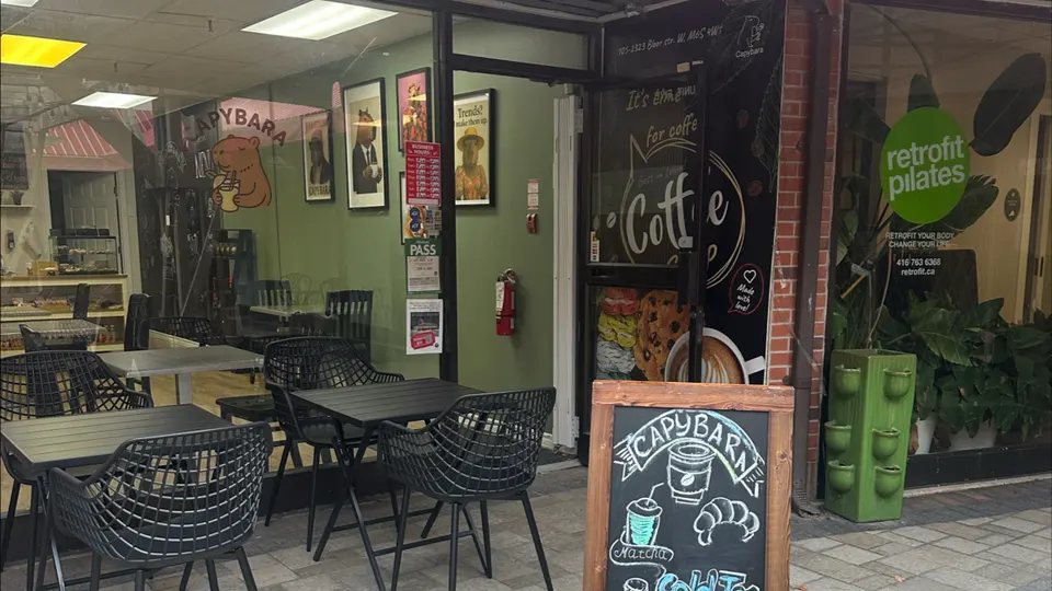 Exterior view of Capybara cafe showing outdoor black metal tables and chairs, a green interior wall with framed capybara art, and a wooden sandwich board sign in the foreground.