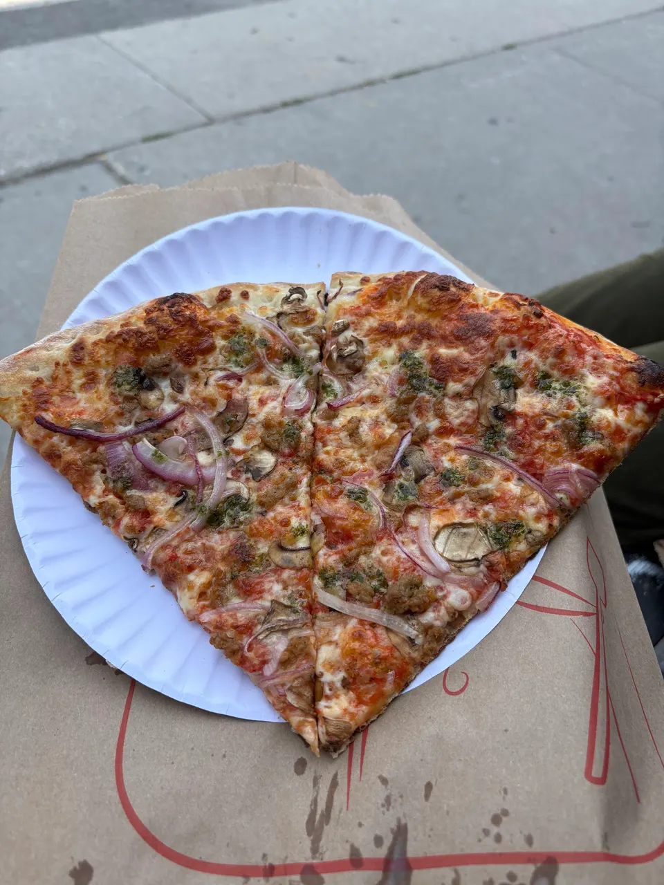 Two slices of thin-crust pizza topped with red onions, mushrooms, sausage, and pesto, served on a white paper plate over a brown paper bag.