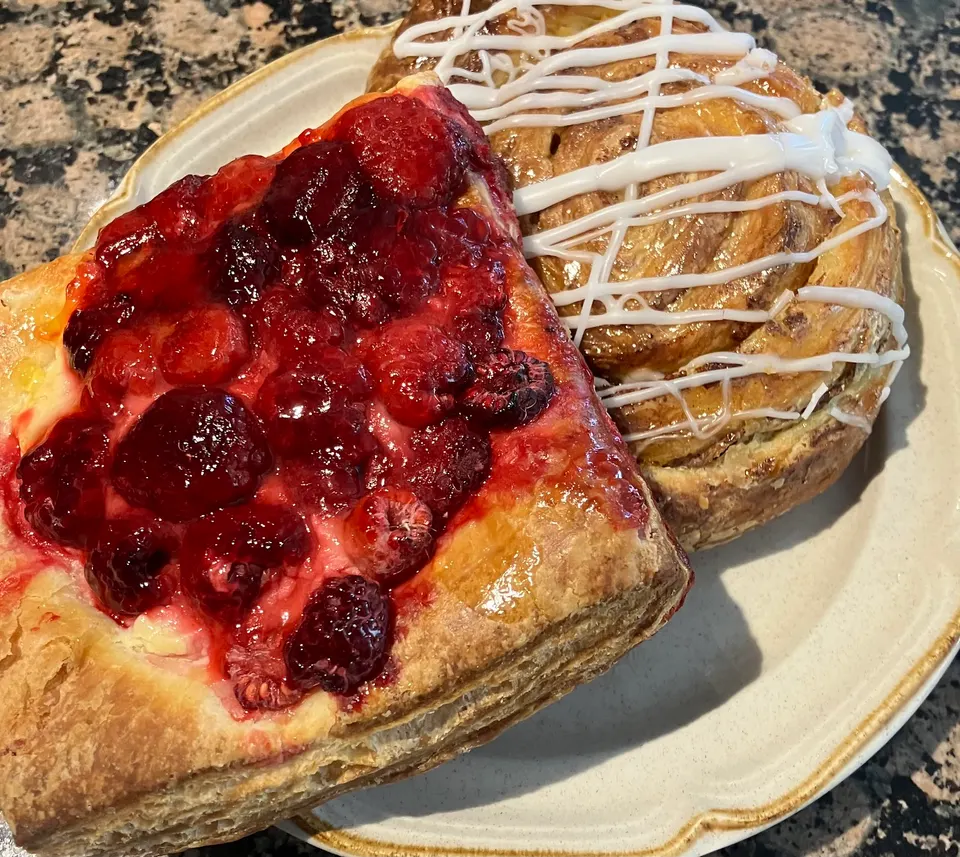 A close-up of two gluten-free pastries on a plate: a square tart topped with glossy red berries and a round cinnamon roll drizzled with white icing.
