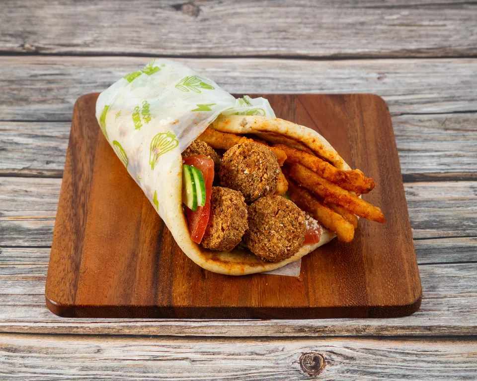 A Greek falafel pita wrap filled with golden falafel balls, seasoned french fries, sliced tomatoes, and cucumber, served on a wooden cutting board over a rustic wooden table.