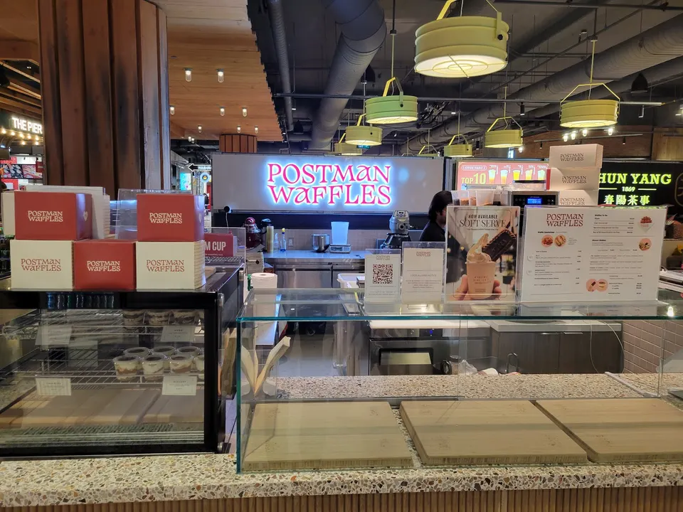 A brightly lit interior view of the Postman Waffles counter, featuring a display case with waffle boxes, a menu stand, and a large illuminated 'POSTMAN WAFFLES' sign in the background. Several light green pendant lights hang from the ceiling.