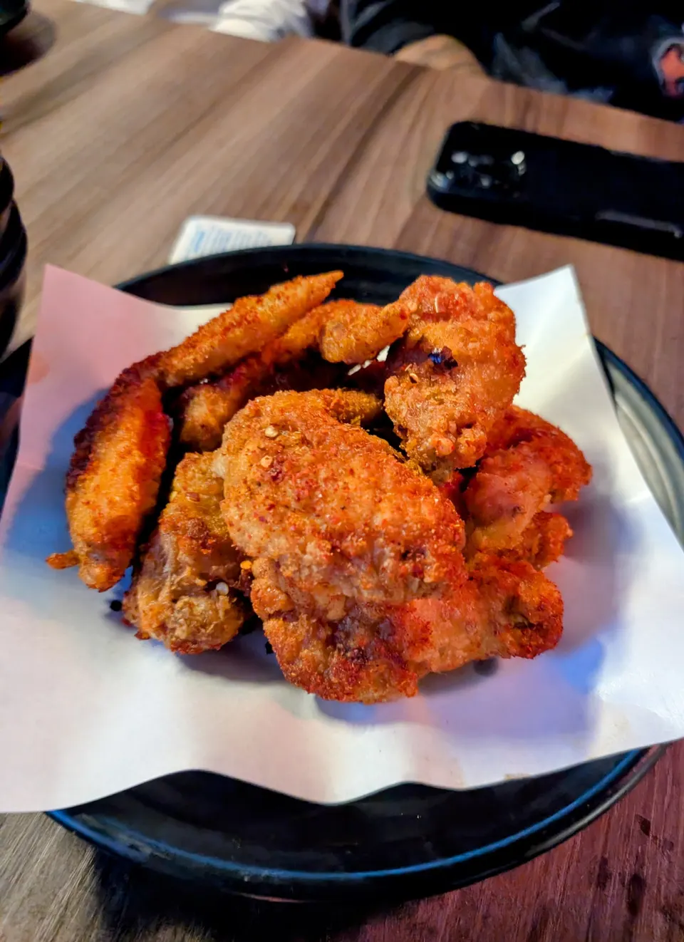 A plate of crispy, golden-brown fried chicken wings seasoned with red chili powder, served on white paper over a black plate on a wooden table.