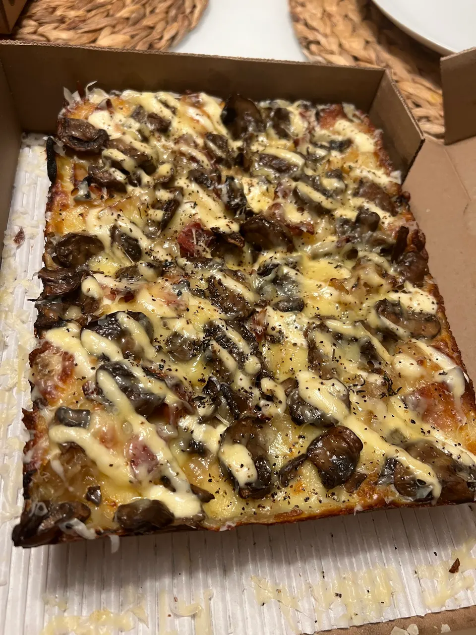 A rectangular Detroit-style deep dish pizza in a cardboard box, topped with melted cheese, caramelized onions, mushrooms, and a creamy white drizzle.
