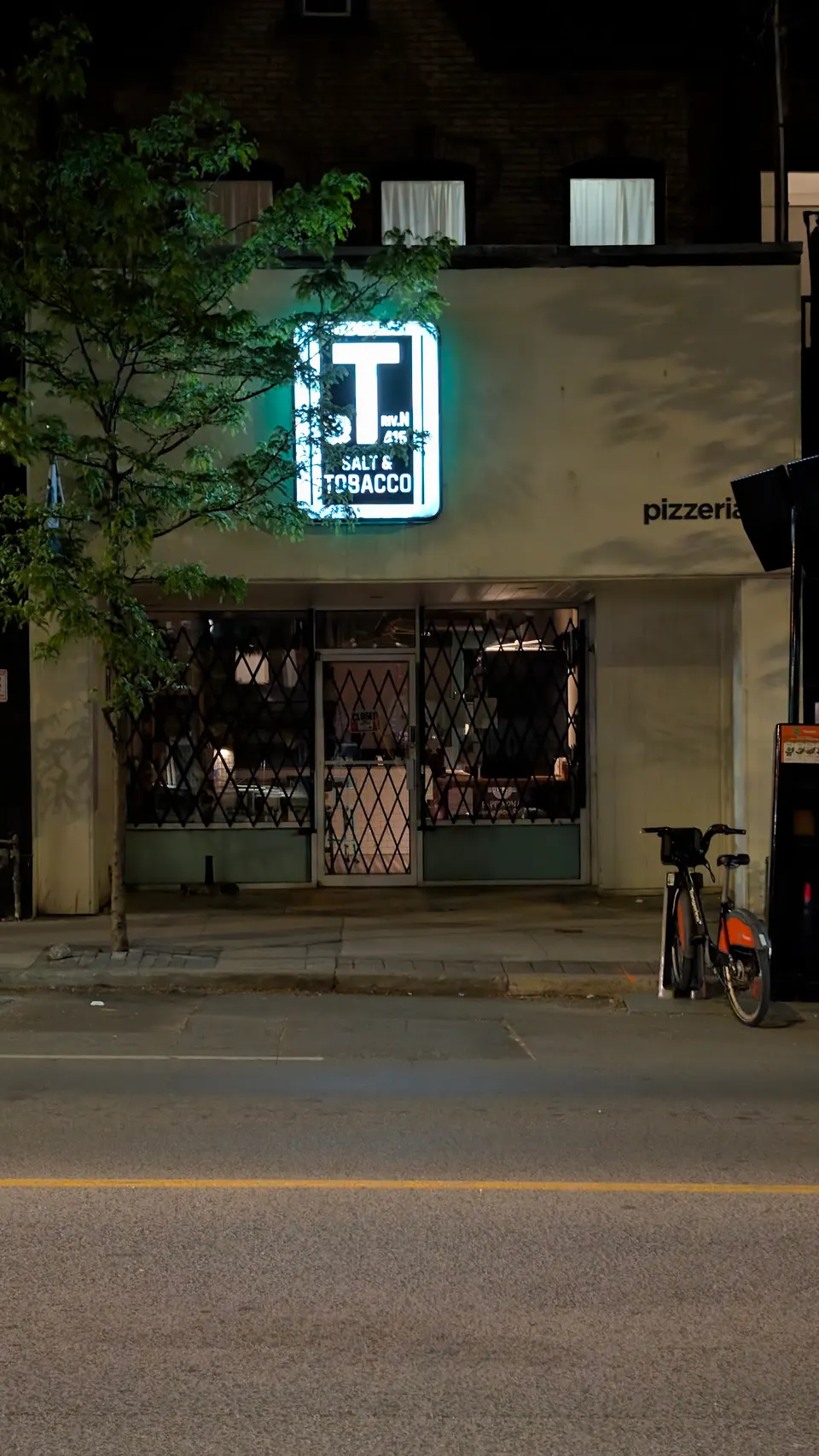 The storefront of Salt & Tobacco Pizzeria at night, featuring a glowing neon sign with the restaurant's name, a metal security gate over the windows, and a bicycle parked on the sidewalk.