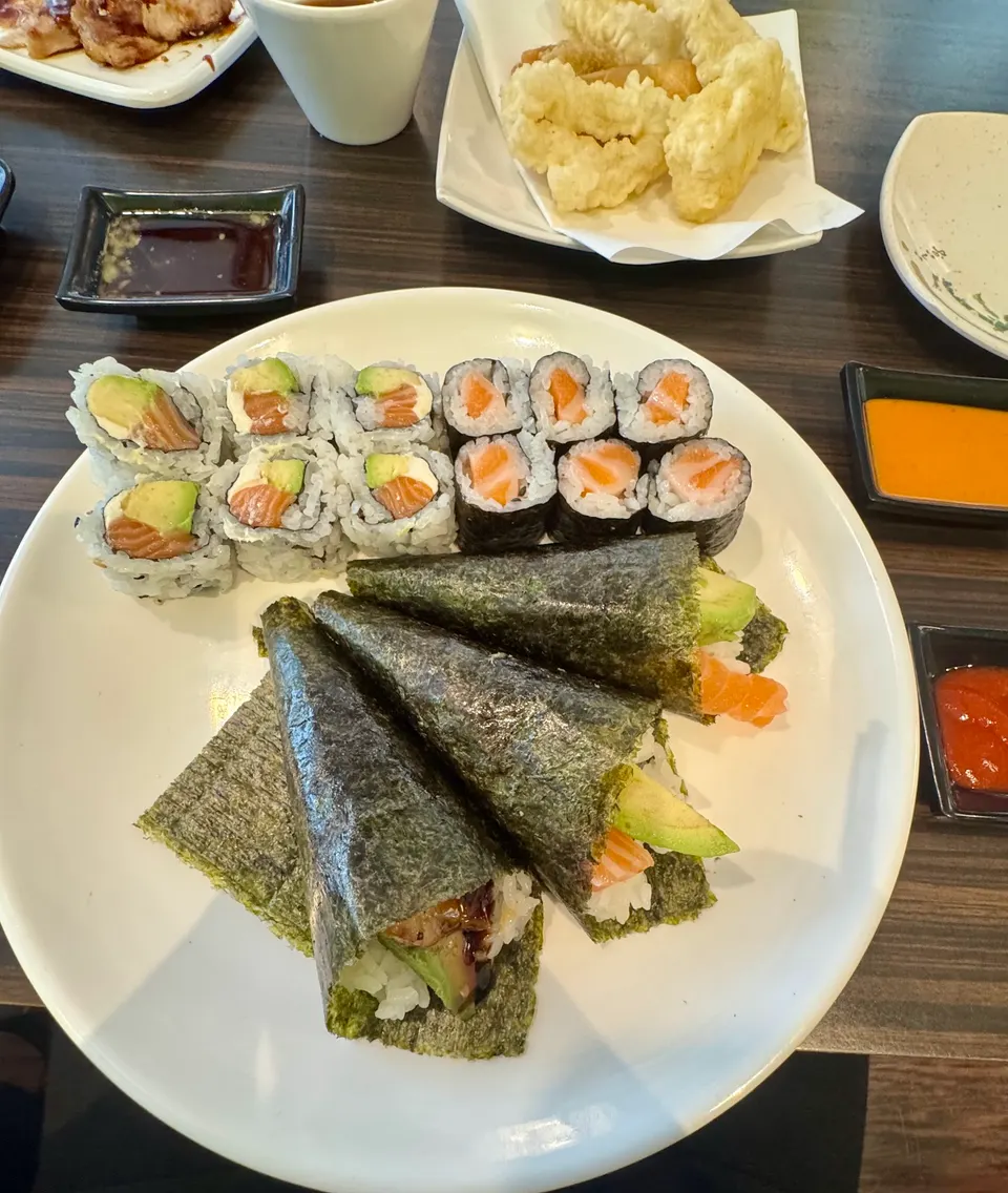 A large white plate filled with an assortment of sushi, including salmon avocado rolls, salmon maki, and three hand rolls wrapped in nori, served alongside tempura and dipping sauces on a dark wooden table.