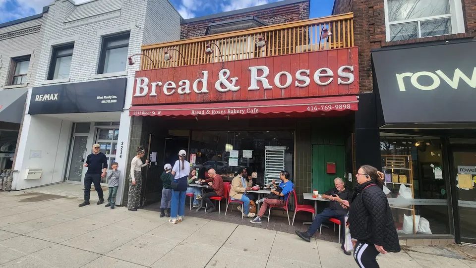 Exterior view of Bread & Roses Bakery Cafe on Bloor Street, featuring a red wooden storefront sign, outdoor seating with patrons, and pedestrians on the sidewalk.