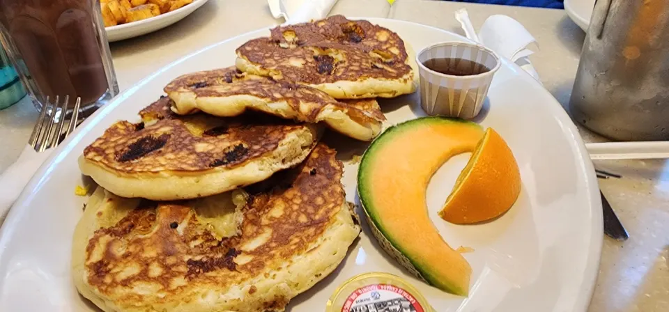 A plate of golden-brown pancakes served with a slice of cantaloupe, an orange wedge, a small cup of syrup, and butter on a white plate.