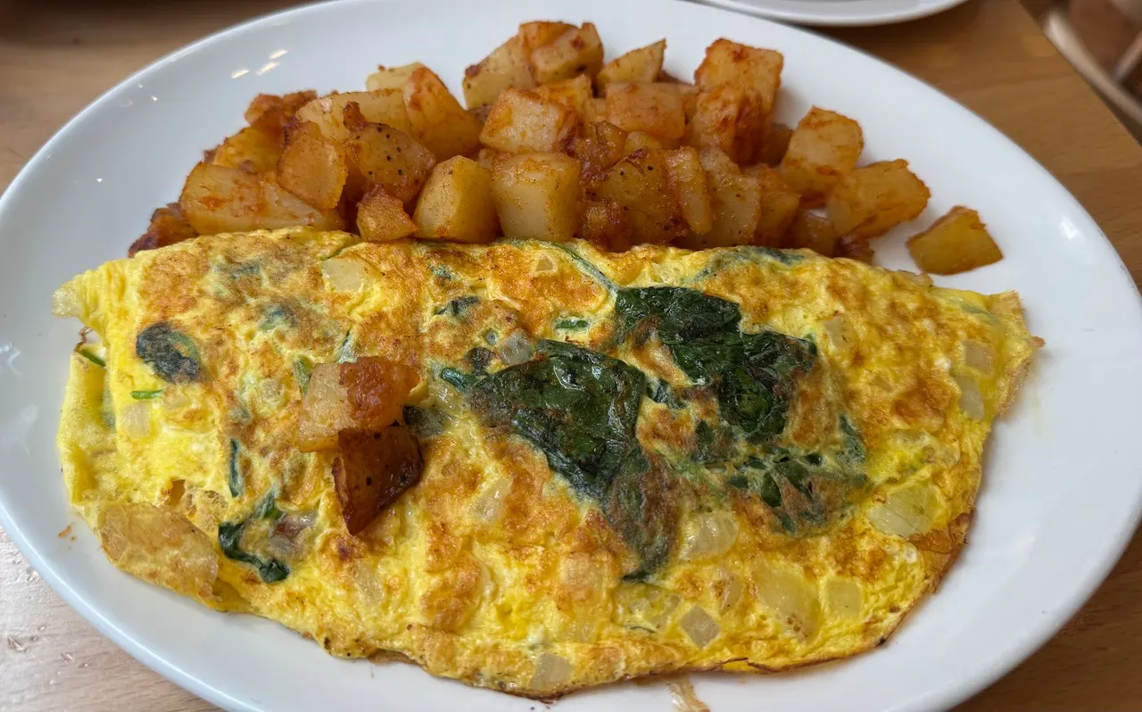 A golden-brown omelette filled with spinach and onions, served on a white plate alongside a generous portion of seasoned, cubed home fries.