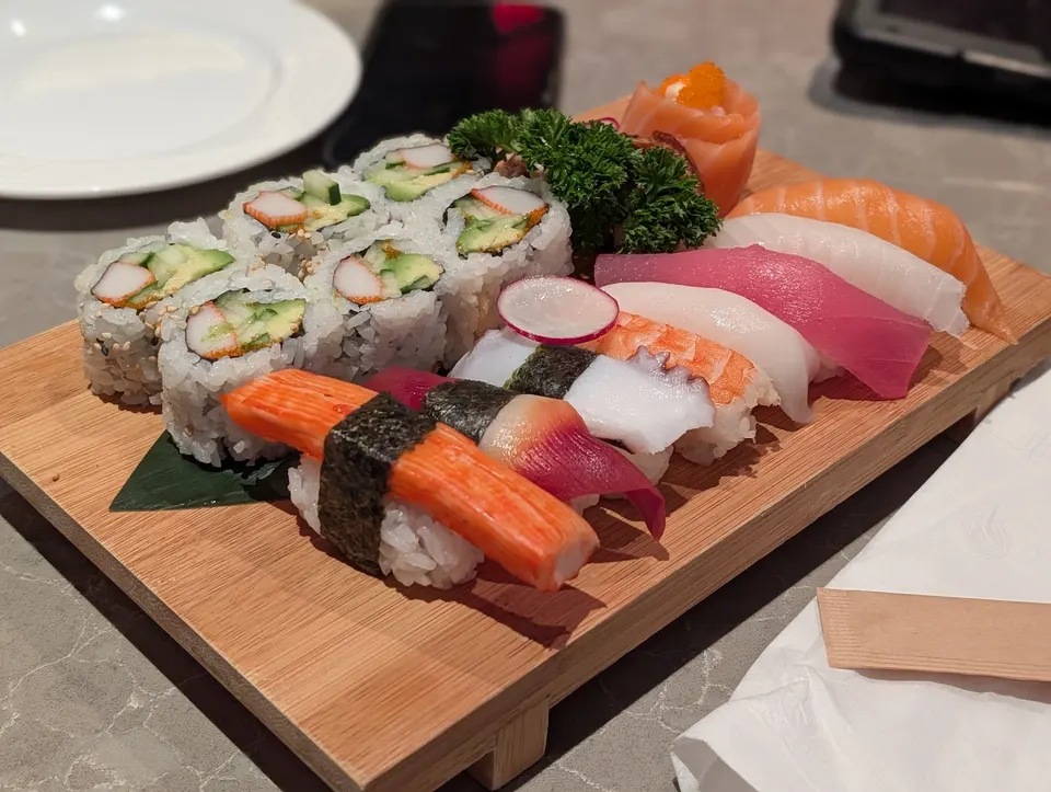 A wooden platter filled with an assortment of sushi, including maki rolls, nigiri with salmon, tuna, shrimp, and crab stick, garnished with parsley and radish slices.