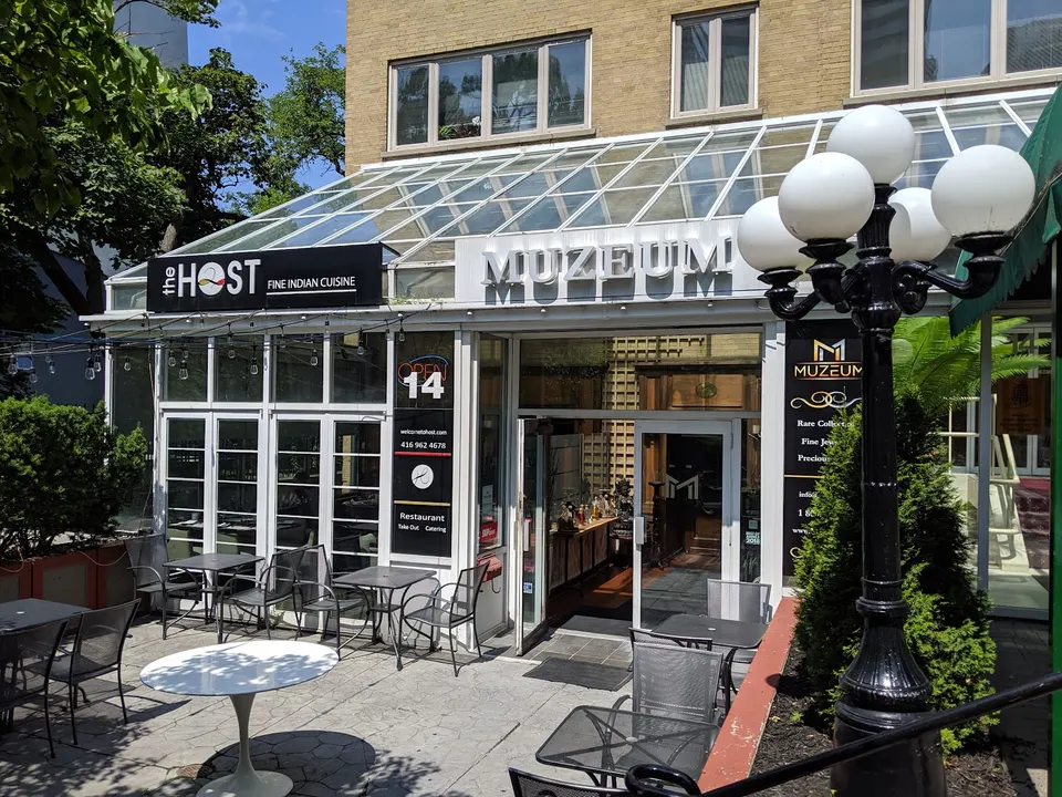The storefront of The Host Fine Indian Cuisine in Toronto, featuring an outdoor patio with metal tables and chairs under a glass atrium roof next to Muzeum.