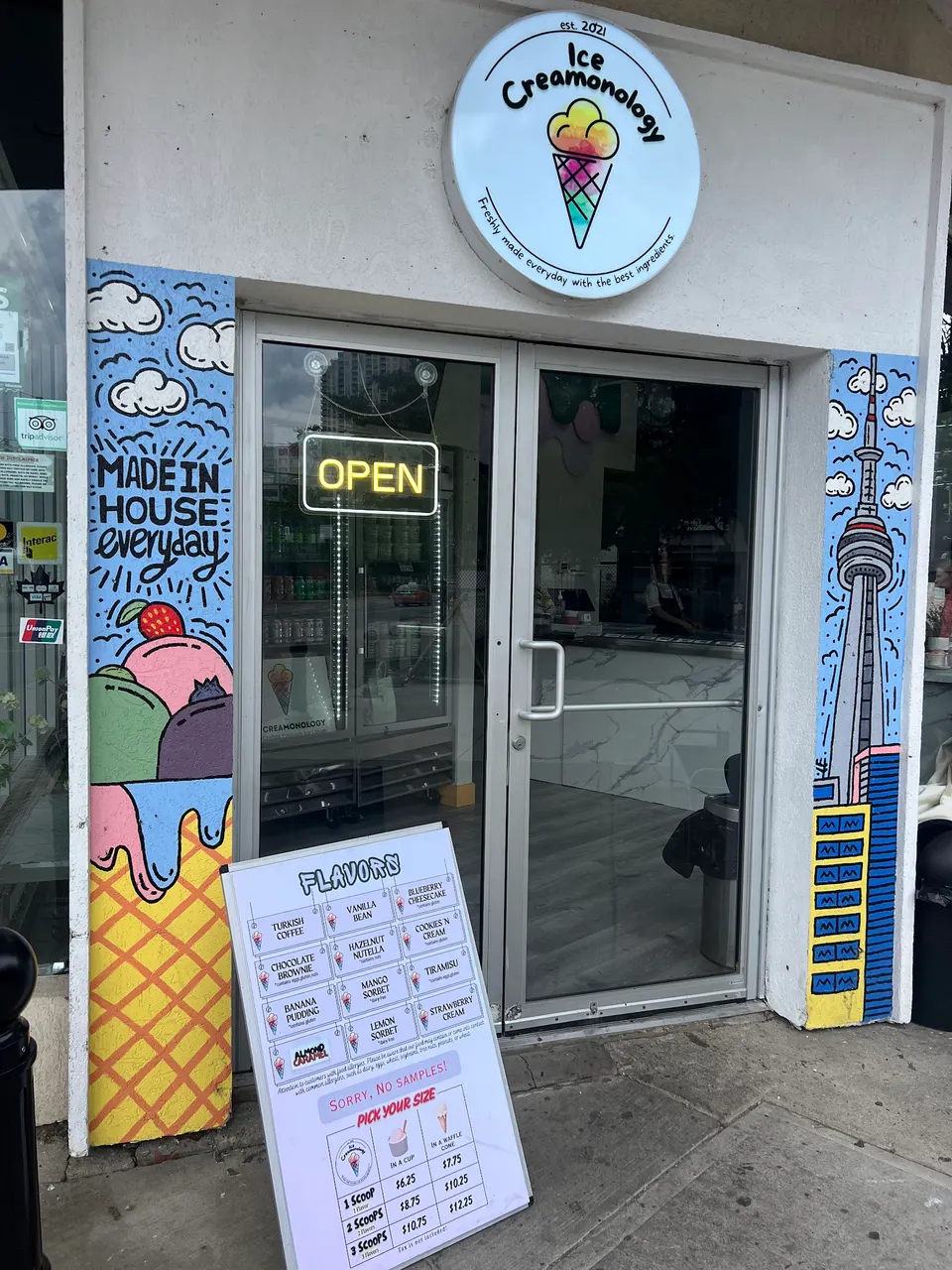 The storefront of Ice Creamonology featuring a round logo sign, hand-painted murals on the door frame, an open sign, and a sandwich board menu displaying flavors and prices.