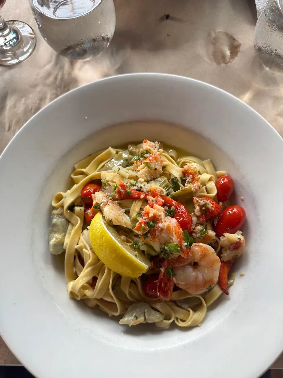 A white bowl filled with fettuccine pasta topped with lobster meat, shrimp, cherry tomatoes, artichoke hearts, and a fresh lemon wedge.