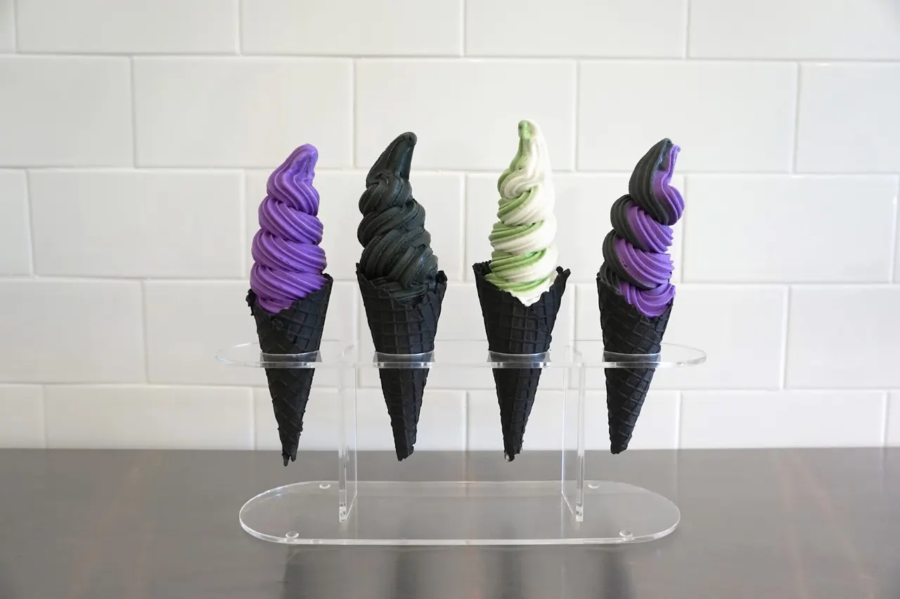 Four soft-serve ice cream cones in black waffle cones, featuring purple, black, green and white, and purple-black swirl flavors, displayed in a clear acrylic stand against a white tiled wall.