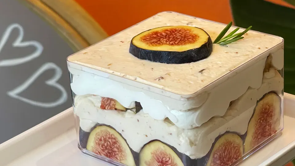 A square clear plastic container filled with a layered fig dessert, topped with a fresh fig slice and a rosemary sprig, resting on a white tray.