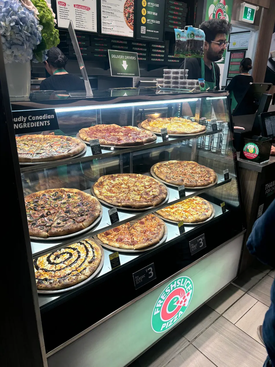 A glass display case at Freshslice Pizza filled with various whole pizzas on metal trays, including pepperoni, vegetarian, and cheese options, with staff visible in the background.