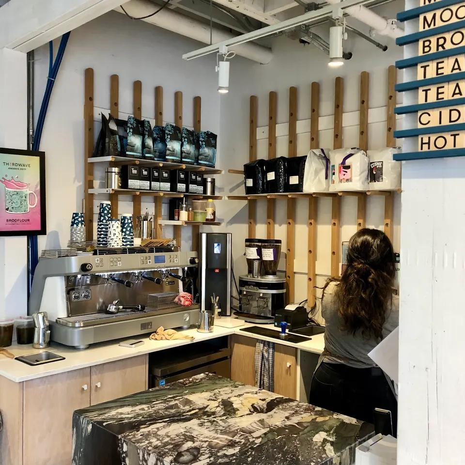 A barista with long dark hair stands behind a wooden counter in a coffee shop, with an espresso machine, coffee grinders, and shelves of coffee beans in the background.