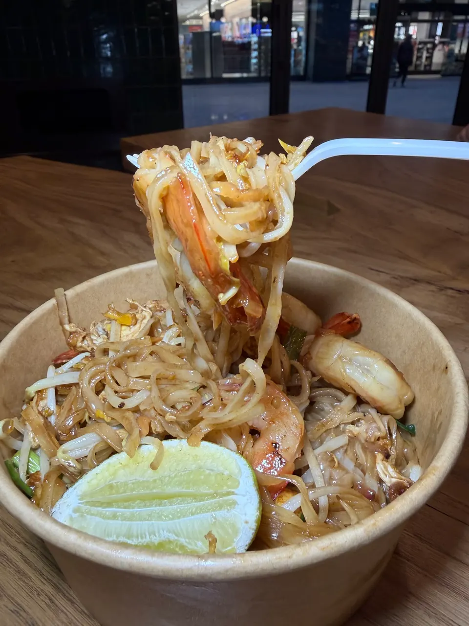 A close-up of a brown paper bowl filled with Pad Thai, featuring flat rice noodles, shrimp, bean sprouts, and scrambled egg. A white plastic fork lifts a portion of noodles and shrimp from the bowl. A lime wedge rests on the rim of the bowl. The background is blurred, showing an indoor setting with 