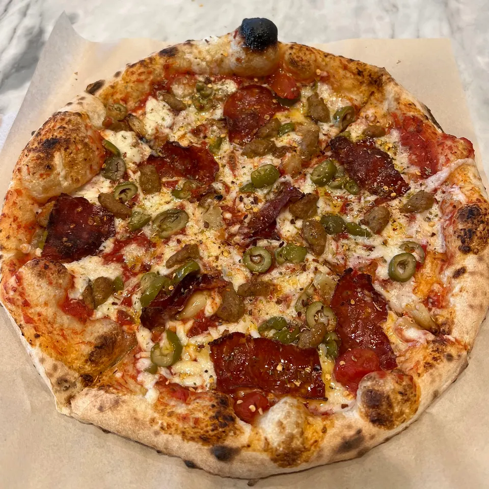 A freshly baked personal pizza with a charred crust, topped with melted cheese, tomato sauce, pepperoni, sausage, sliced green olives, and red pepper flakes on parchment paper.