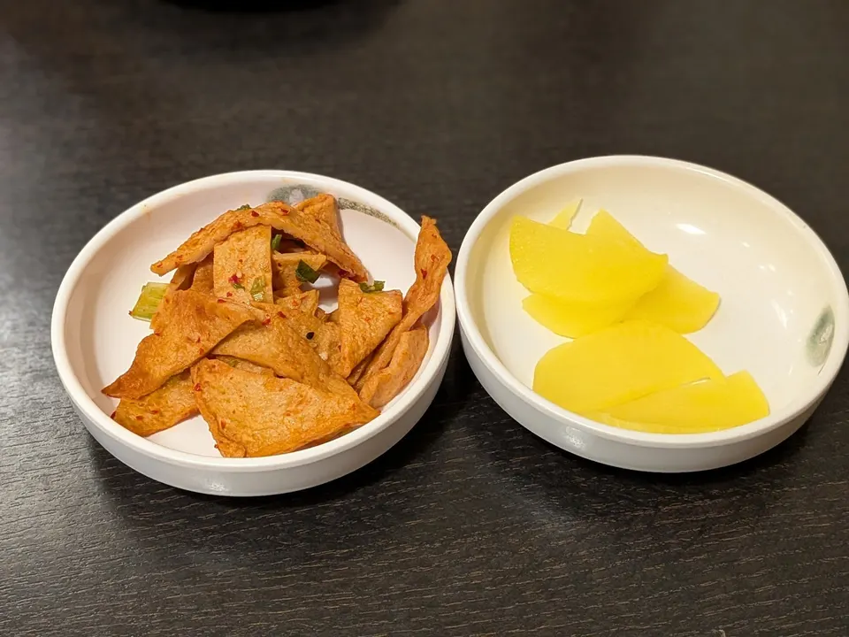 Two small white side dishes on a dark table, one containing seasoned fish cake strips and the other containing yellow pickled radish slices.