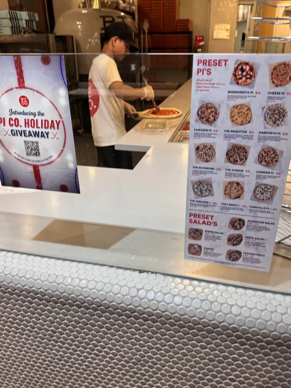 A worker prepares a pizza behind a glass partition at Pi Co., with a holiday giveaway poster on the left and a printed menu of preset pies and salads on the right.