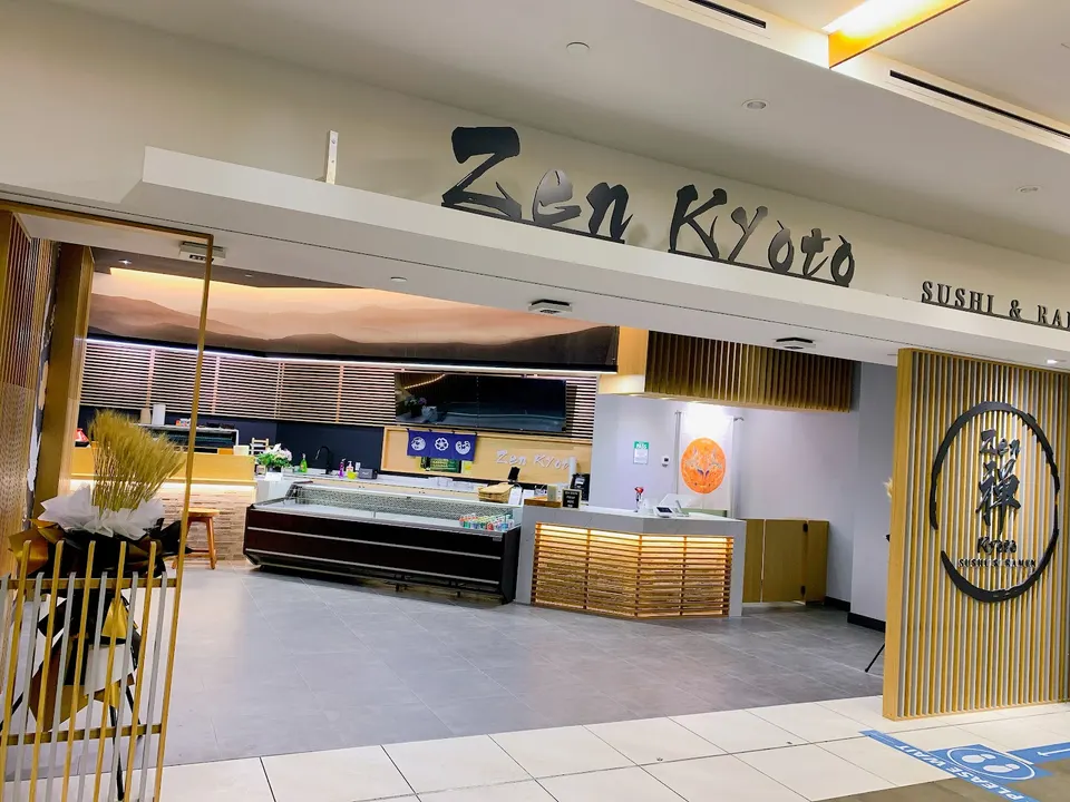 The storefront of Zen Kyoto Sushi & Ramen, featuring a wide open entrance, a light wood counter, a refrigerated display case, and a large sign reading Zen Kyoto.